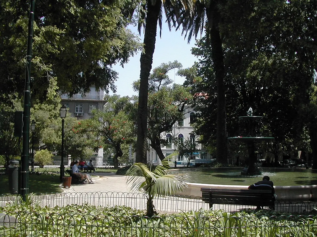Jardim com palmeiras, lago com repuxo e bancos, rodeado de árvores e edifícios.