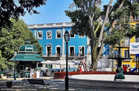 Jardim Alfredo Keil em Lisboa, com quiosque verde, esplanada, fonte e parque infantil junto a edifício azul