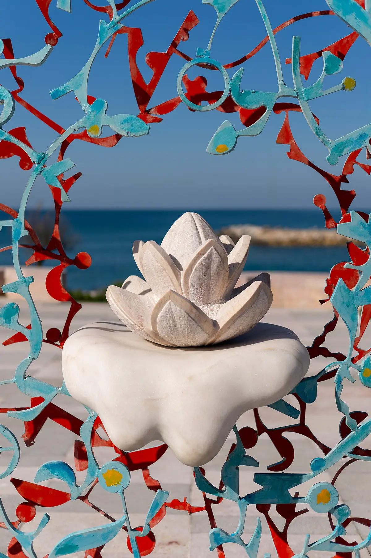 Escultura de flor de lótem em mármore branco enquadrada por gradeamento colorido, com o mar ao fundo