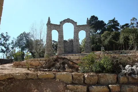 Ruína decorativa em arcos de pedra sobre muros de pedra, rodeada de árvores, sob céu azul