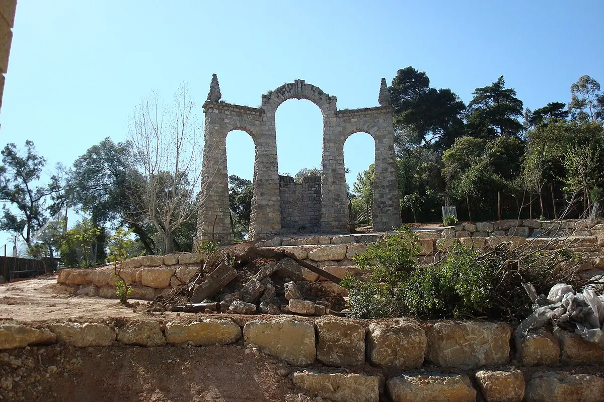 Ruína decorativa em arcos de pedra sobre muros de pedra, rodeada de árvores, sob céu azul