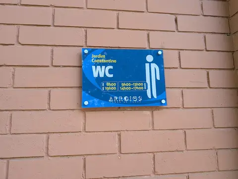 Placa azul com horário dos WC públicos do Jardim Constantino, em parede de tijolo