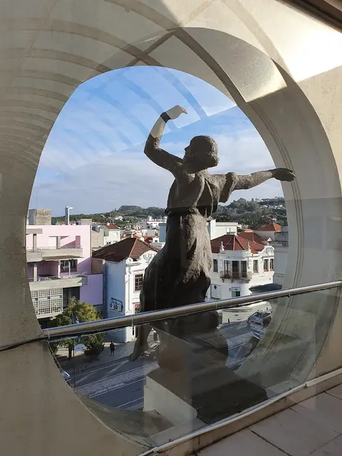 Escultura vista através de janela circular do museu, com vista sobre a vila e casas.