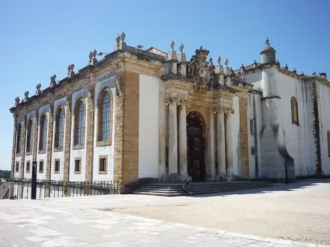 Fachada exterior barroca da Biblioteca Joanina, em Coimbra, com portal e escadaria sob céu azul.