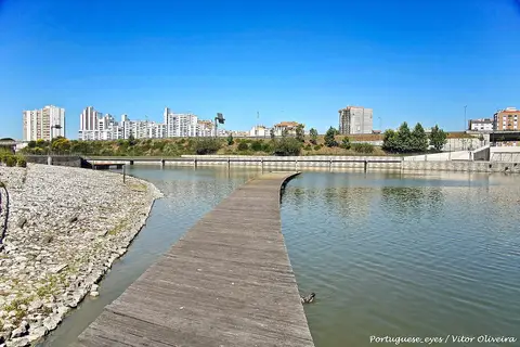 Passadiço de madeira sobre o lago do Parque Oeste, com edifícios ao fundo e céu azul