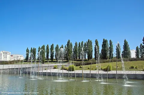 Lago com repuxos de água no Parque Oeste, com fila de choupos e relvado ao fundo