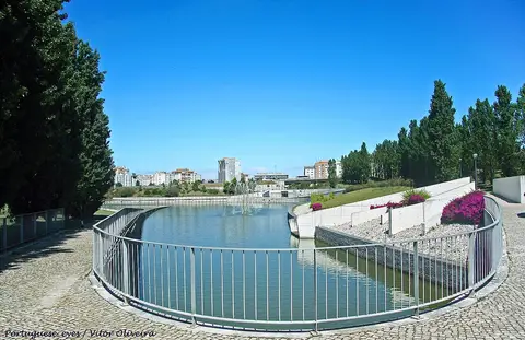 Lago com repuxo no Parque Oeste, em Lisboa, rodeado por guarda metálica e árvores