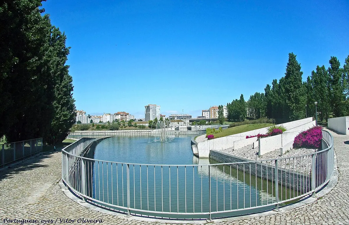 Lago com repuxo no Parque Oeste, em Lisboa, rodeado por guarda metálica e árvores