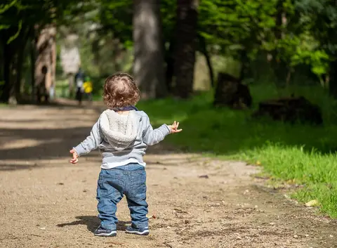 Criança pequena de costas a caminhar num carreiro de terra ladeado de árvores num parque