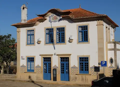 Edifício branco com janelas e portas azuis, azulejos e brasão na fachada, em Arco de Baúlhe.