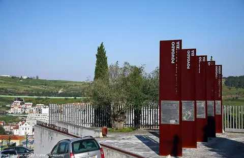 Painéis informativos vermelhos à entrada do Povoado Pré-Histórico de Leceia, com oliveira e vista sobre a vila.