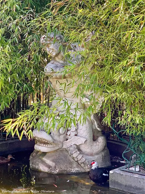 Estátua de pedra com golfinho num lago rodeado de bambus, com patos na água, no Parque Silva Porto