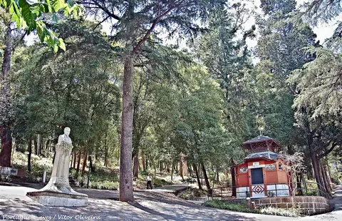 Parque arborizado com estátua em pedra e quiosque de madeira vermelho junto a caminho.