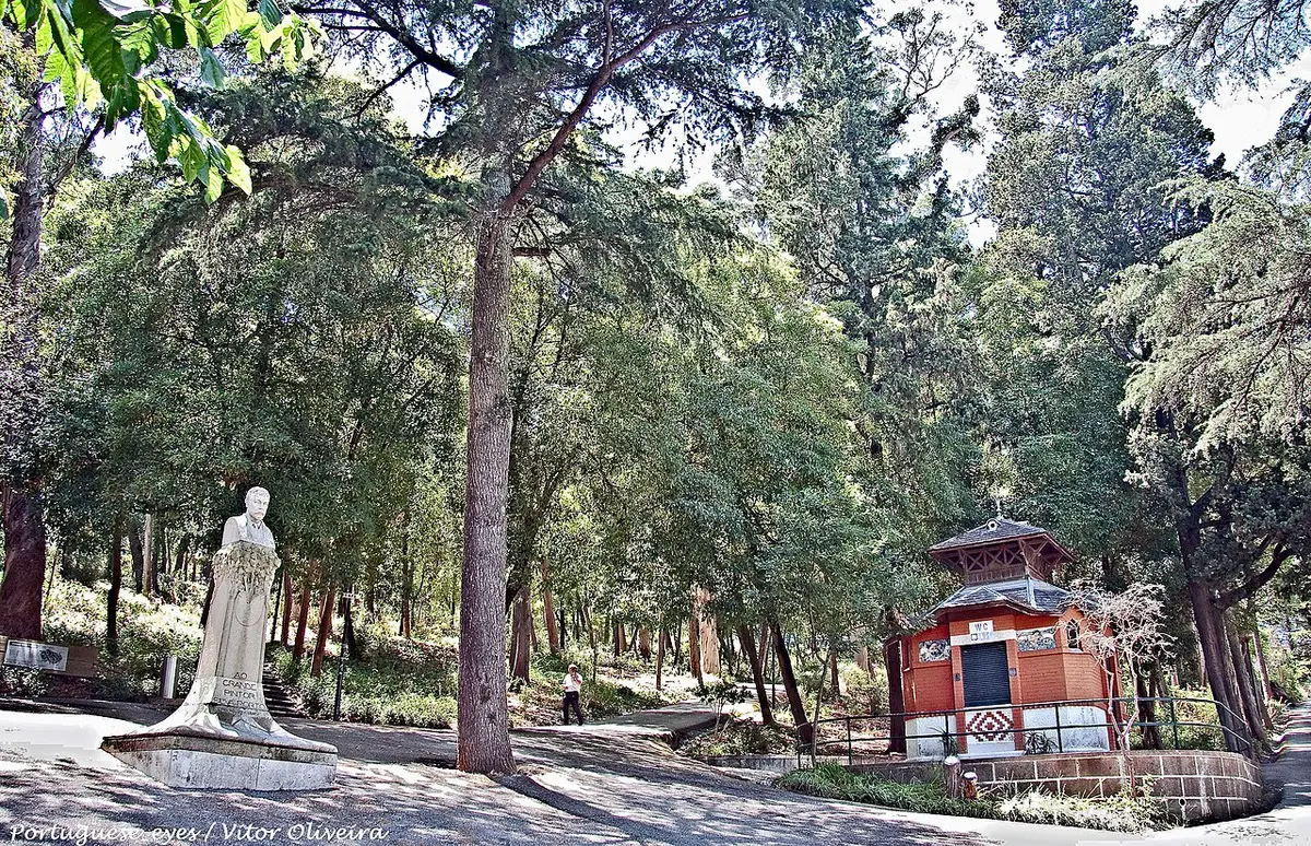 Parque arborizado com estátua em pedra e quiosque de madeira vermelho junto a caminho.