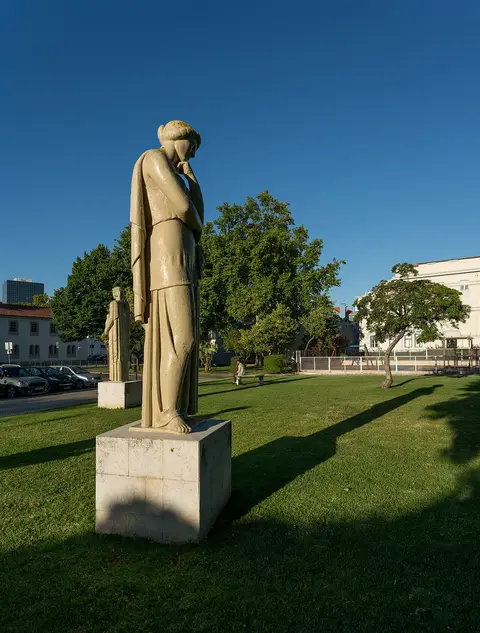 Esculturas de pedra sobre plintos no relvado do Jardim Irmã Lúcia, com árvores e céu azul.