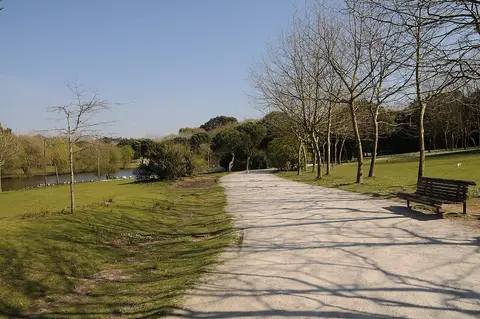 Caminho de saibro num parque urbano, com bancos, relva e lago ao fundo