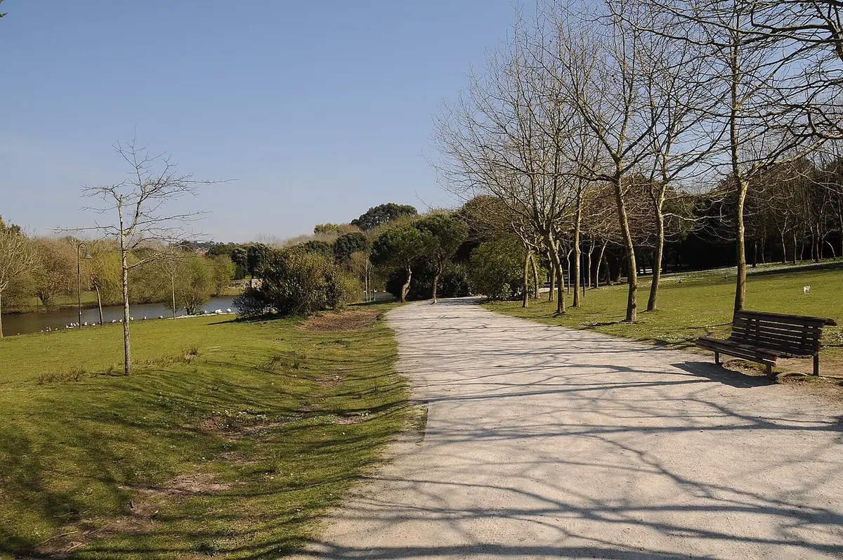 Caminho de saibro num parque urbano, com bancos, relva e lago ao fundo