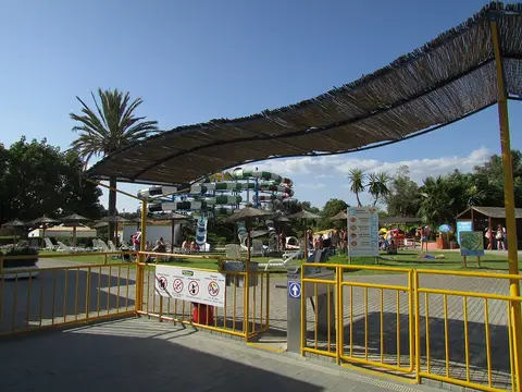 Entrada do parque aquático Aqualand com gradeamento amarelo, toldo de colmo e escorregas ao fundo
