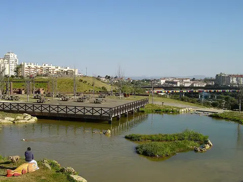 Lago e deque de madeira no Parque da Bela Vista, com zona de merendas e prédios ao fundo