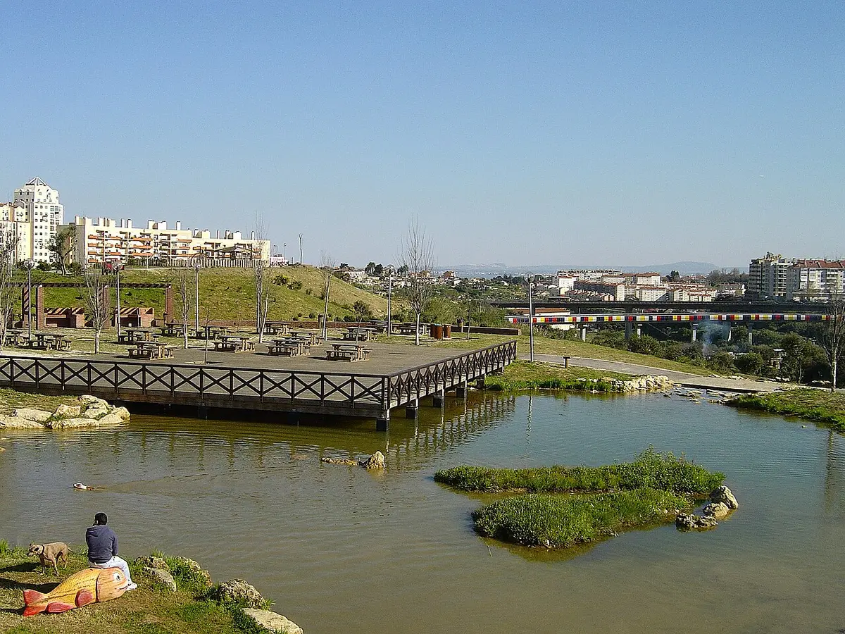 Lago e deque de madeira no Parque da Bela Vista, com zona de merendas e prédios ao fundo