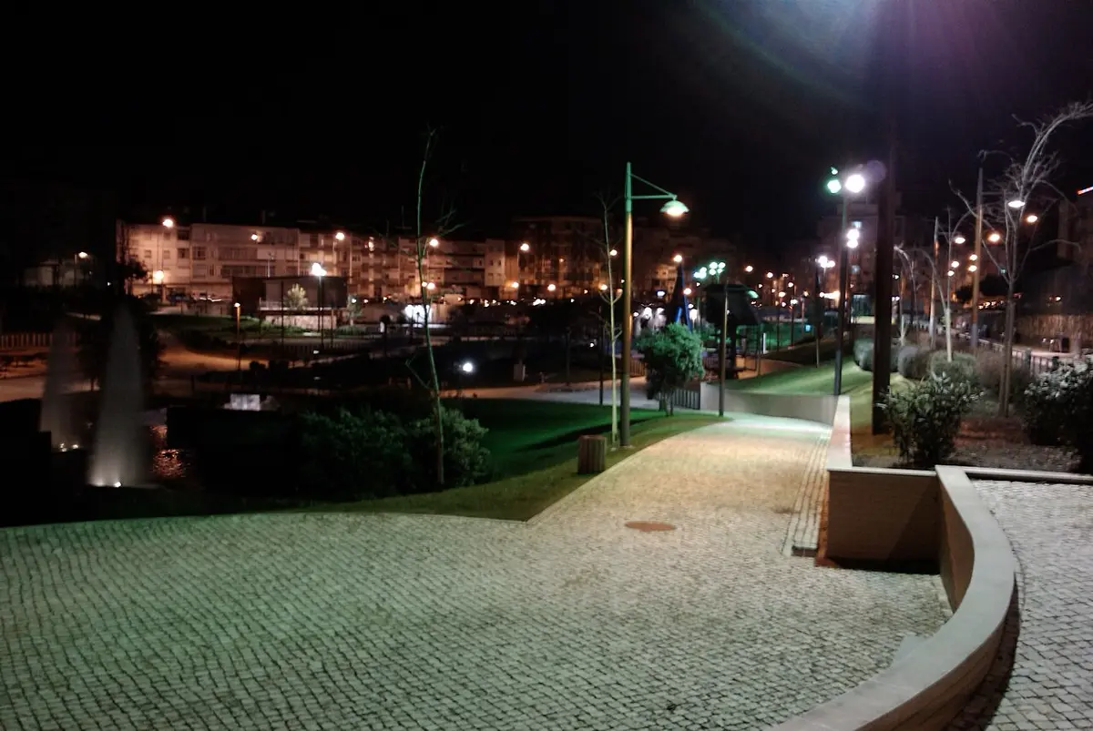 Parque urbano à noite, com passeio em calçada, repuxo de água iluminado e candeeiros acesos.