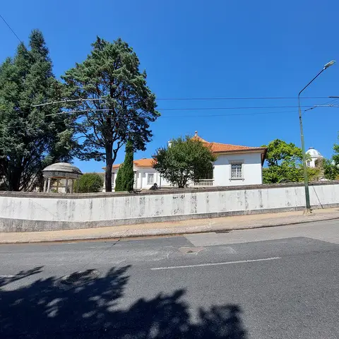 Vista exterior da Casa Museu Bissaya Barreto, com muro branco, jardim arborizado e coreto, sob céu azul.
