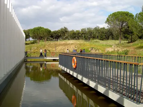 Passadiço sobre espelho de água junto ao edifício do Fluviário de Mora, com árvores ao fundo