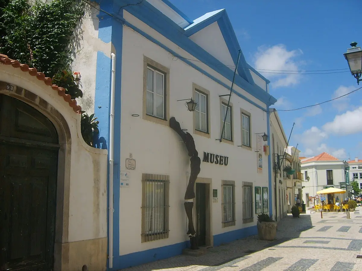 Fachada branca e azul do Museu da Lourinhã, com escultura na parede e rua calcetada