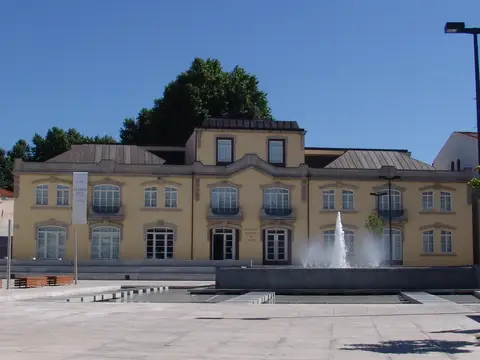 Fachada amarela da Biblioteca Municipal de Chaves com praça e repuxo de água em primeiro plano