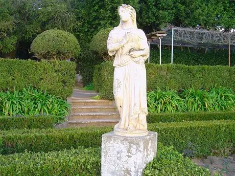 Estátua de pedra sobre pedestal num jardim com sebes aparadas e escadaria ao fundo