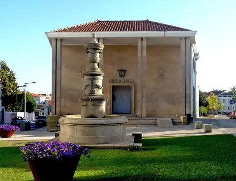 Edifício da biblioteca municipal com fonte de pedra e canteiro de flores roxas em primeiro plano