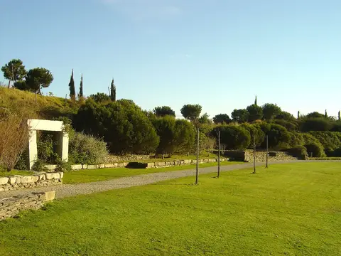 Relvado e caminho de terra no Parque da Paz, com pórtico de pedra e árvores ao fundo