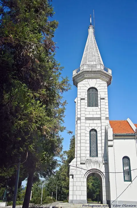 Torre branca com coruchéu do Santuário de Nossa Senhora dos Milagres, rodeada de árvores, sob céu azul