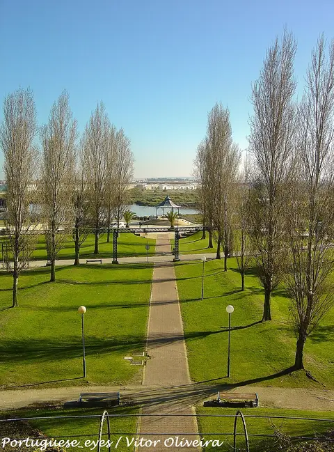 Vista do parque municipal com caminho central ladeado de relva, árvores e coreto junto à água.
