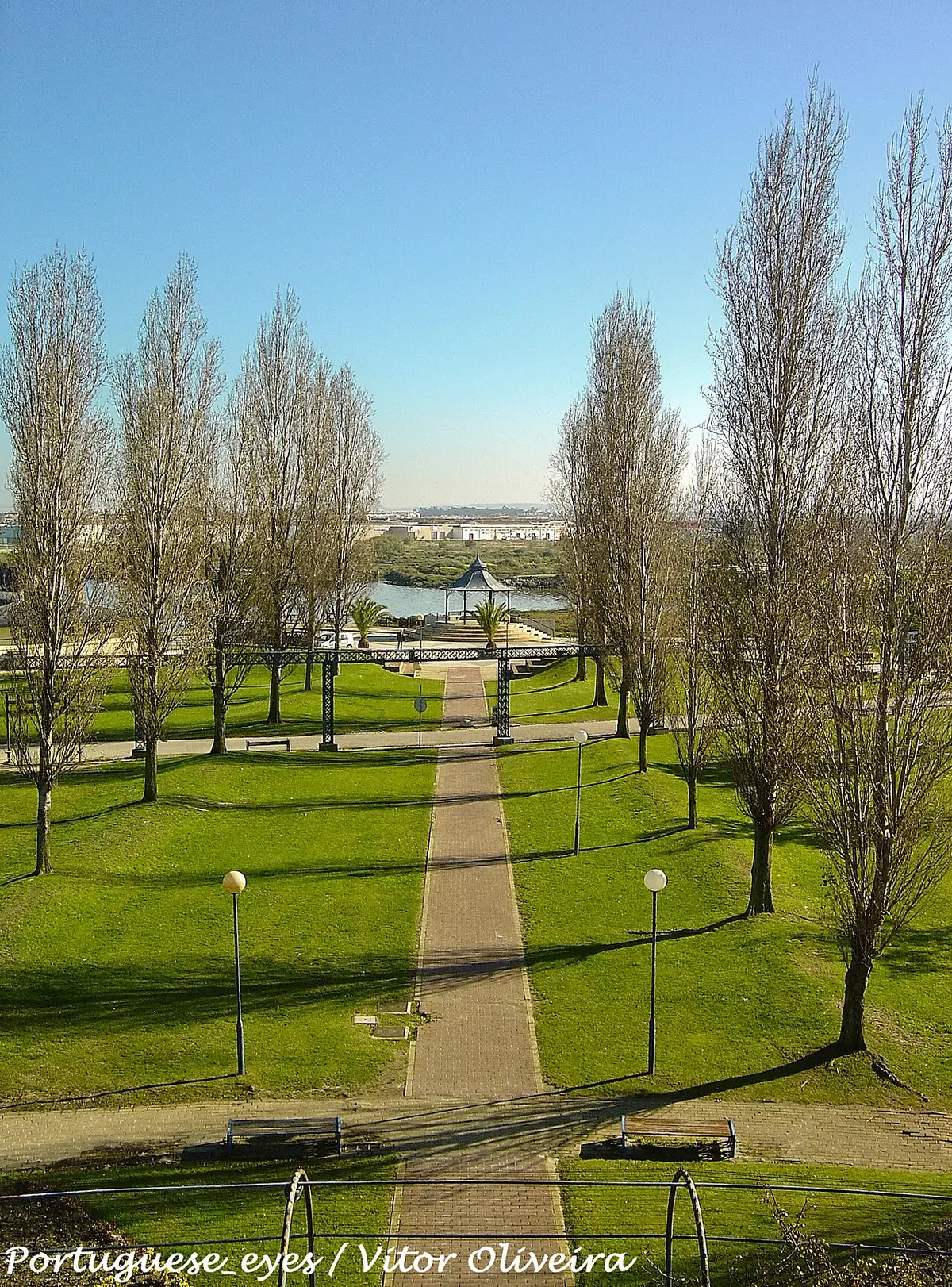 Vista do parque municipal com caminho central ladeado de relva, árvores e coreto junto à água.