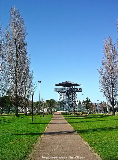 Torre metálica de observação ao fundo de um caminho empedrado, com relvados e árvores no parque.