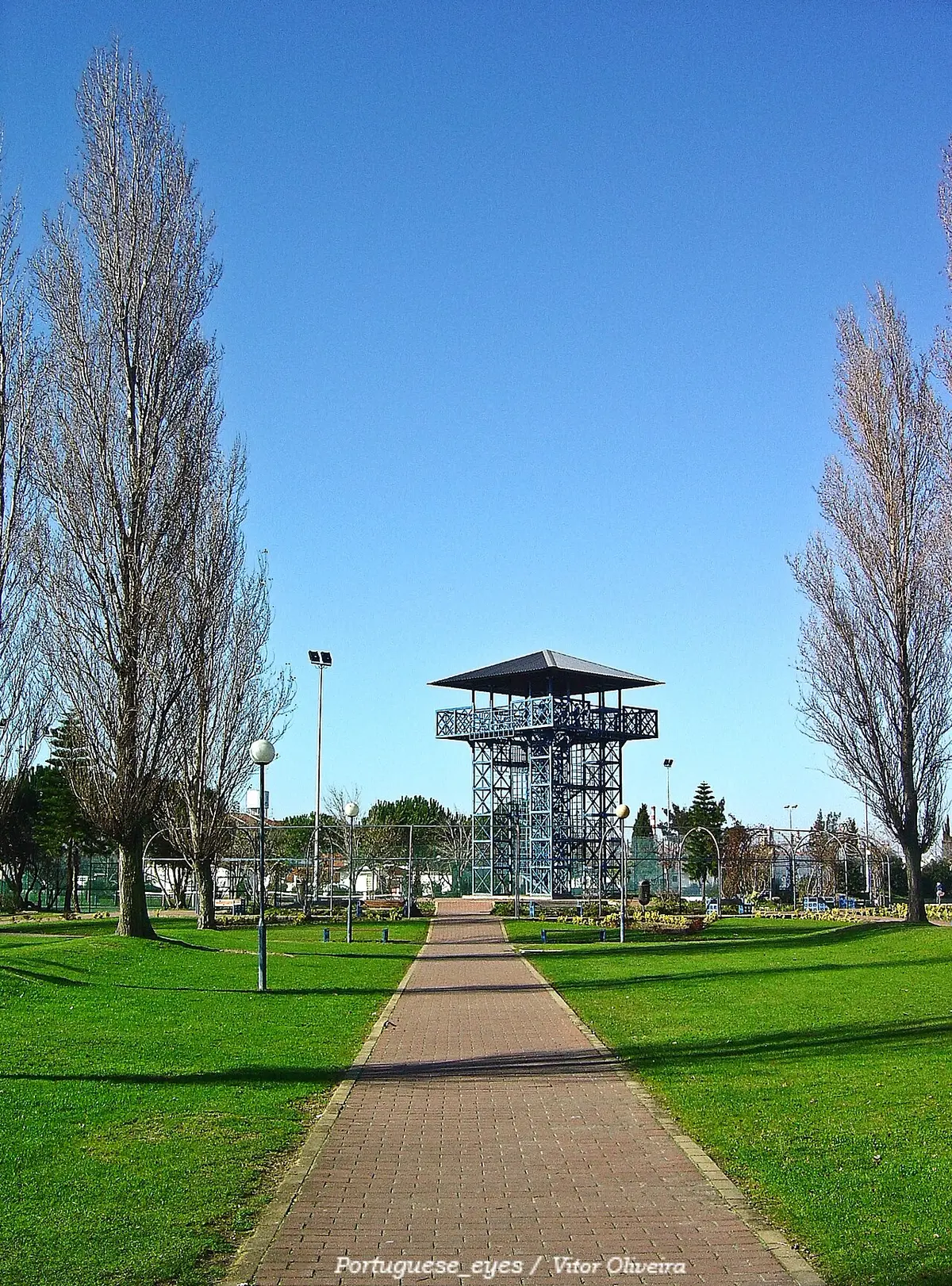 Torre metálica de observação ao fundo de um caminho empedrado, com relvados e árvores no parque.