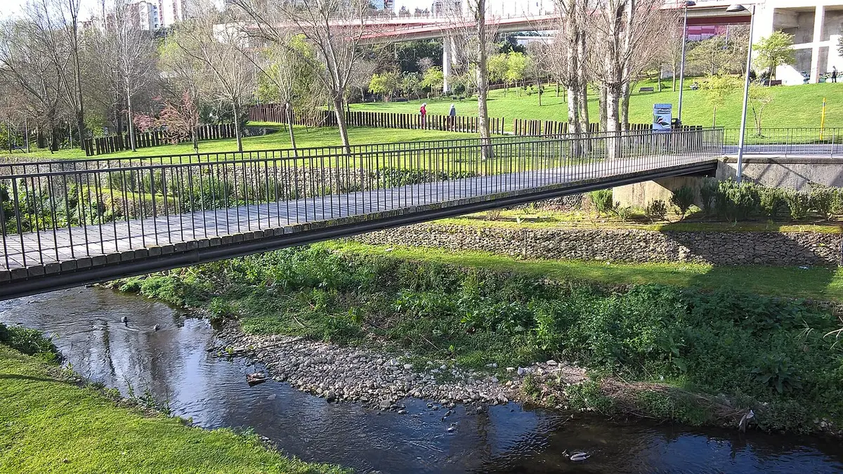 Ponte pedonal sobre ribeiro no Parque Linear Dom Domingos Jardo, com relvados e árvores em redor