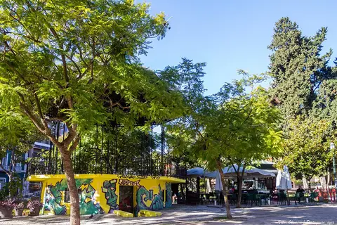 Jardim com árvores e quiosque amarelo decorado com grafítis, junto a esplanada