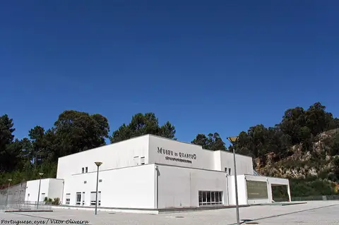 Edifício branco do Museu do Quartzo, com letreiro na fachada, rodeado de pinhal sob céu azul.