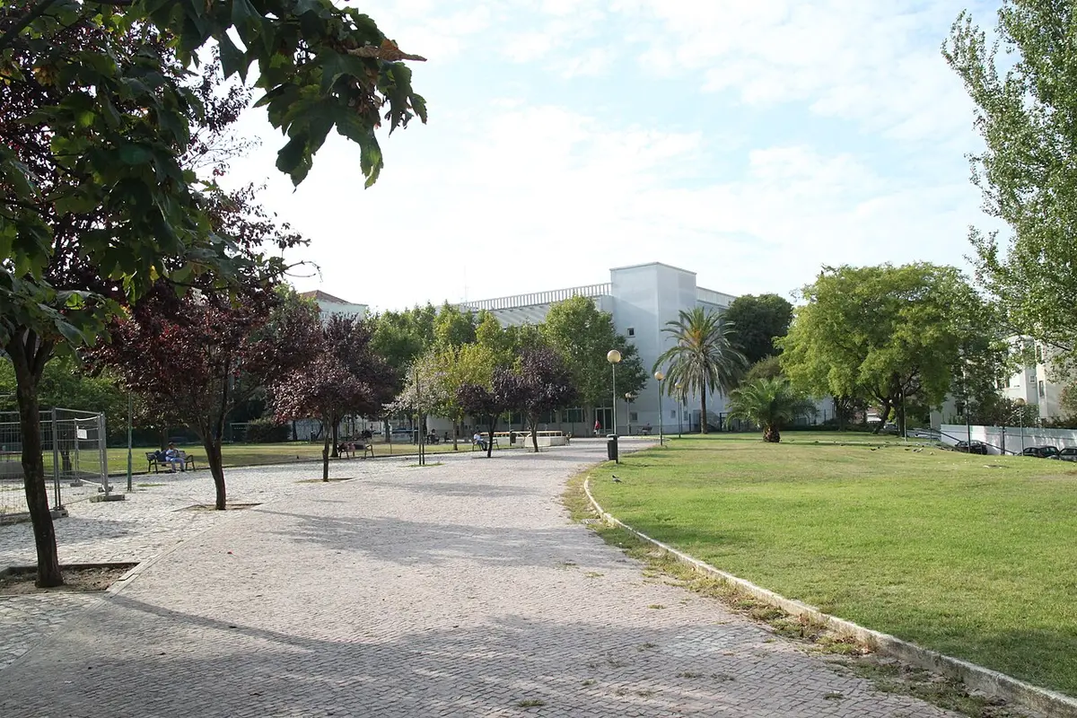 Jardim com caminho calcetado, relvado e árvores, com edifício branco ao fundo