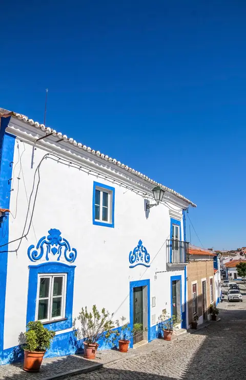 Casa caiada de branco com frisos azuis e vasos de plantas numa rua calcetada de Messejana.