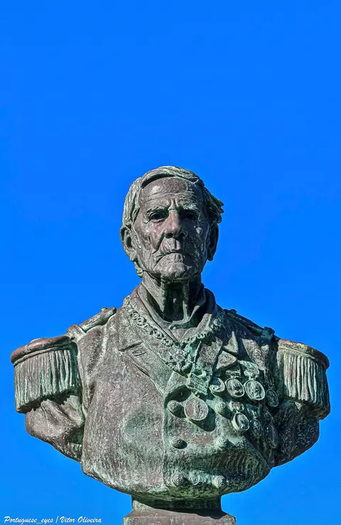 Busto em bronze do Patrão Joaquim Lopes, fardado com medalhas, contra céu azul, em Paço de Arcos
