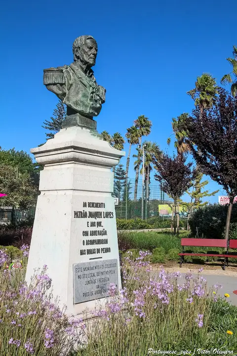 Busto do Patrão Joaquim Lopes sobre pedestal de pedra, rodeado de flores e palmeiras no jardim de Paço de Arcos