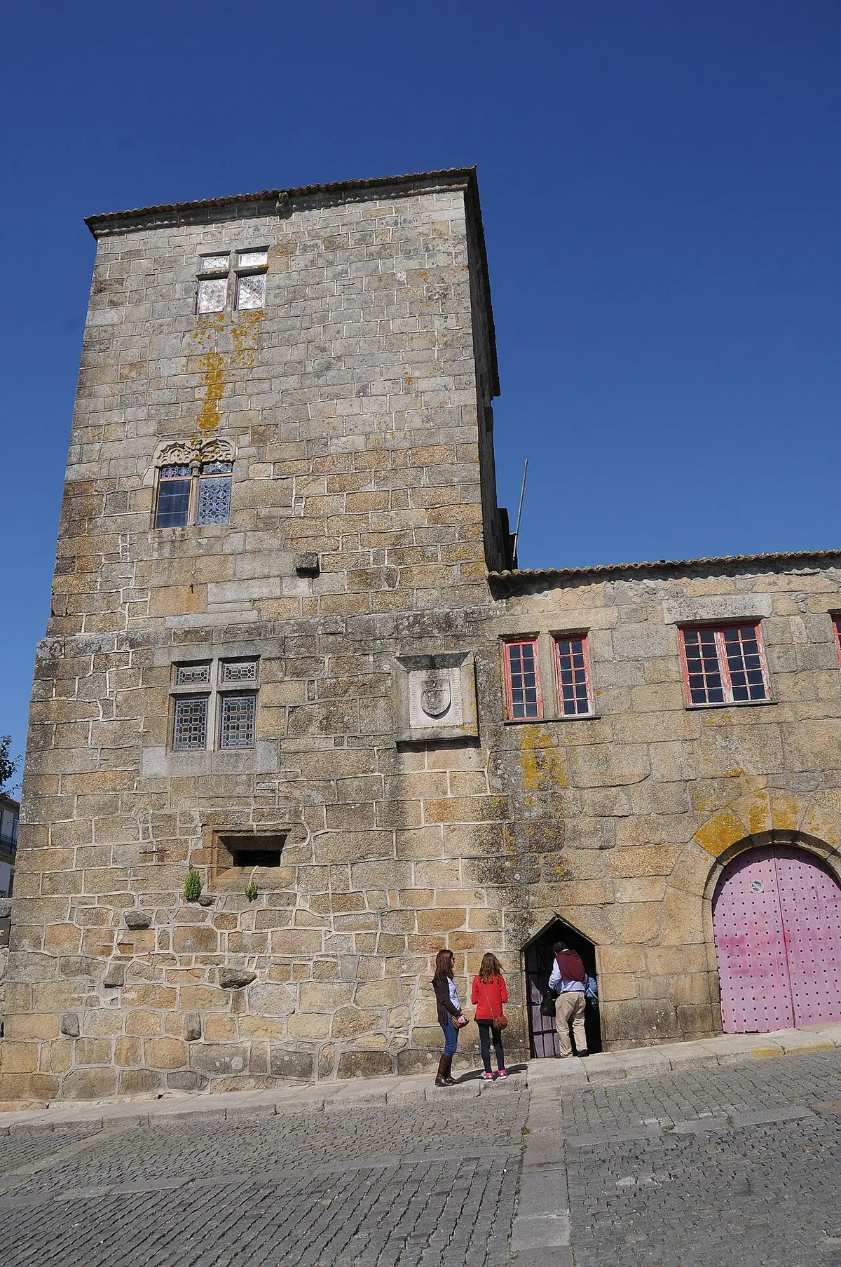 Fachada em granito do Solar dos Pinheiros, com torre medieval, janelas manuelinas e portão vermelho.
