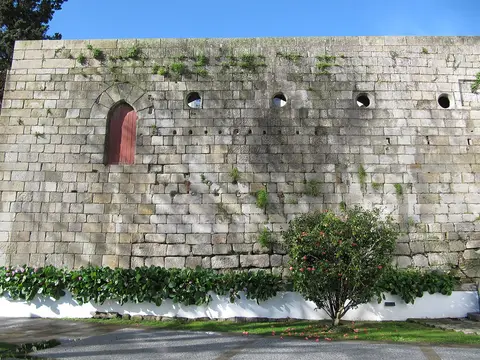 Muro alto de pedra com porta em arco de madeira, vegetação rasteira e arbusto florido à frente.