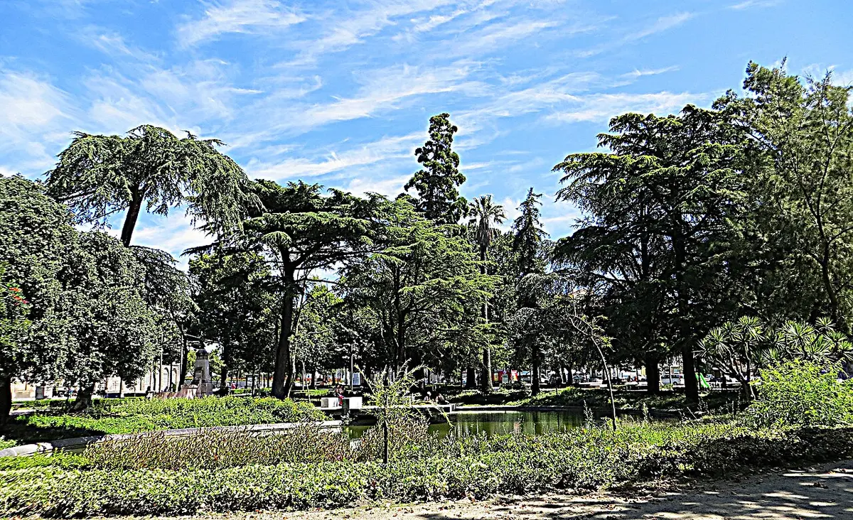 Jardim com árvores altas, canteiros e lago sob céu azul