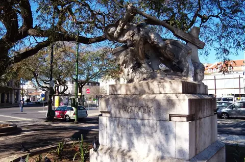 Monumento em pedra com escultura num jardim urbano, rodeado de árvores e rua com automóveis.