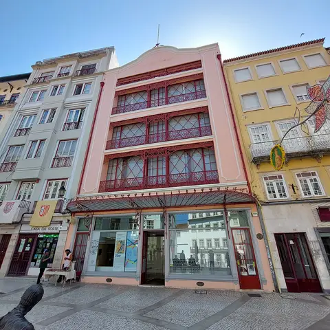 Fachada rosa do Edifício Chiado em Coimbra, com varandas de ferro vermelho e montras ao nível da rua.