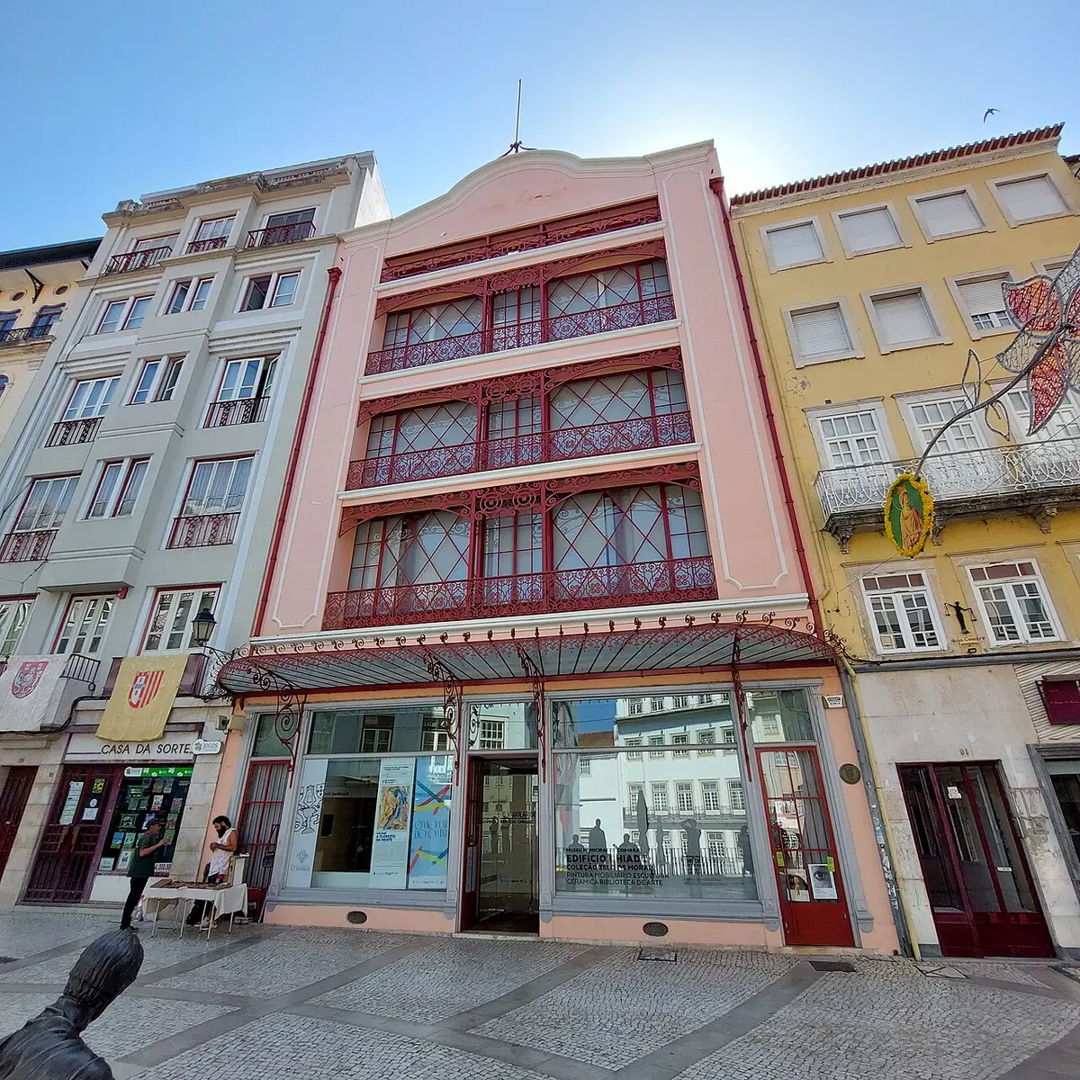 Fachada rosa do Edifício Chiado em Coimbra, com varandas de ferro vermelho e montras ao nível da rua.